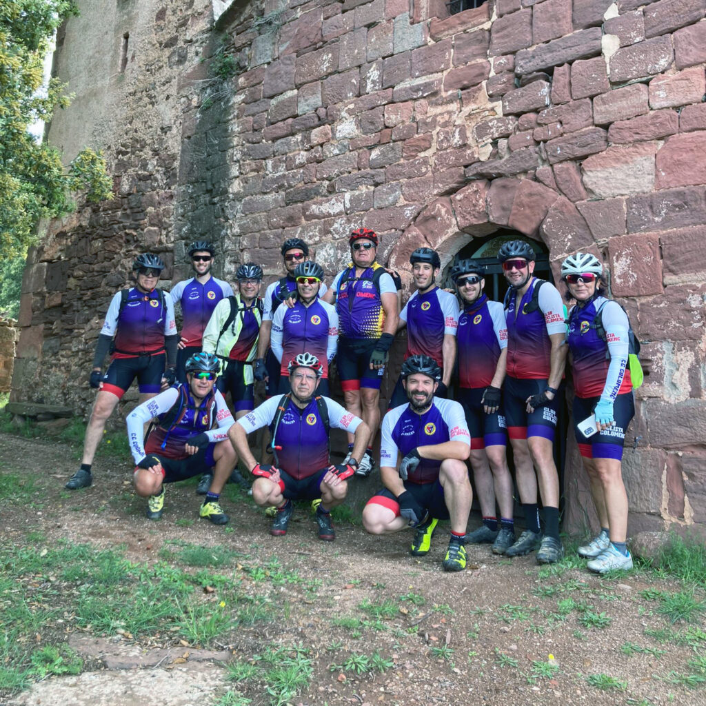 Un grupo de 15 ciclistas posa frente a una antigua construcción de piedra rojiza con una pequeña puerta arqueada. Visten equipaciones a juego en tonos morado, rojo y blanco, con cascos y gafas de sol. Algunos están de pie y otros agachados, con expresiones relajadas. El entorno es natural, con césped y árboles, sugiriendo una ruta ciclista por una zona histórica o rural.