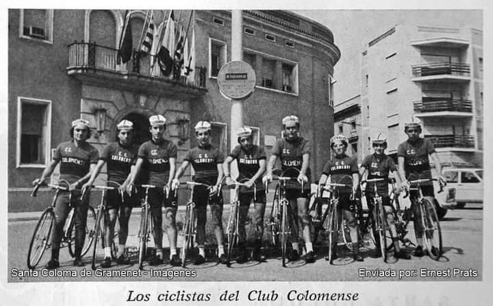 Fotografía en blanco y negro de un grupo de ciclistas del Club Colomense posando con sus bicicletas frente a un edificio con banderas. Llevan uniformes oscuros con gorras y gafas. La imagen muestra una escena clásica de ciclismo en una ciudad.