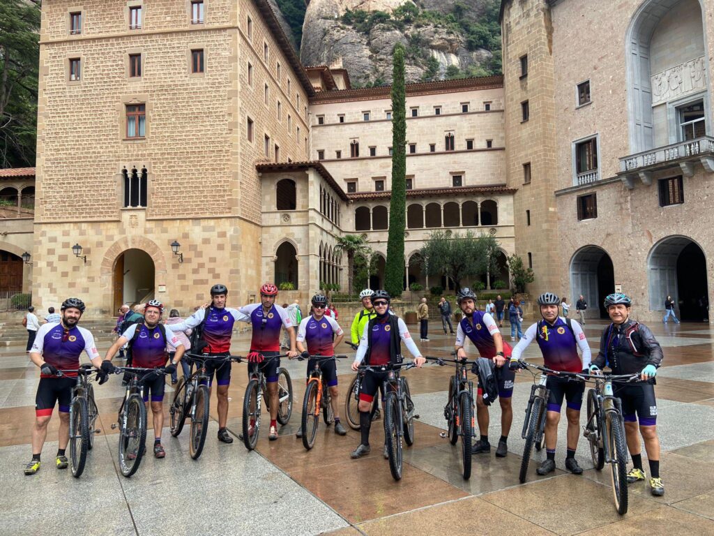 Un grupo de ciclistas posa con sus bicicletas frente a un edificio histórico de piedra con arcos y balcones. Visten equipaciones morado-rojo y algunos llevan cascos y gafas. El suelo está mojado y varias personas caminan alrededor, en un entorno de montaña.